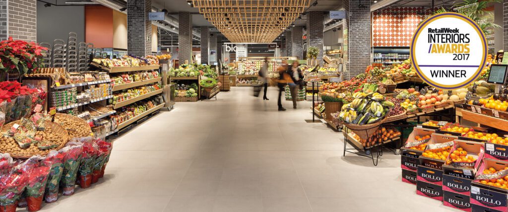 Award-winning grocery store interior with neatly arranged produce, flowers, and packaged goods, featuring a Retail Week Interiors Award 2017 badge.